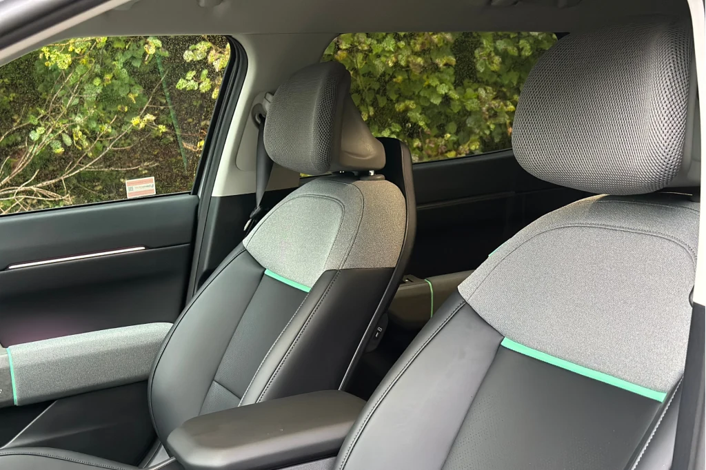 Zagłówki z siatką są wyjątkowo komfortowe. Przednie fotele samochodowe o nowoczesnym designie z szaro-czarną tapicerką, widoczne detale przeszyć oraz fragment drzwi od strony pasażera, w tle zielone liście za szybą.