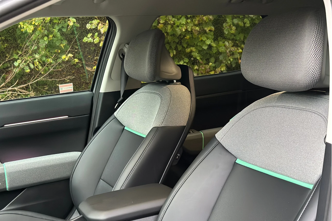 Zagłówki z siatką są wyjątkowo komfortowe. Przednie fotele samochodowe o nowoczesnym designie z szaro-czarną tapicerką, widoczne detale przeszyć oraz fragment drzwi od strony pasażera, w tle zielone liście za szybą.
