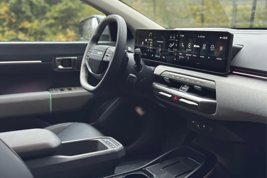 Wnętrze nowej Kii EV4 nie przytłacza nowoczesnością. W zamian za to jest prosto, użytecznie i intuicyjnie. Wnętrze nowoczesnego samochodu z cyfrowym wyświetlaczem na desce rozdzielczej, wielofunkcyjną kierownicą i wygodnymi siedzeniami, widoczne elementy klimatyzacji oraz przestronny podłokietnik.