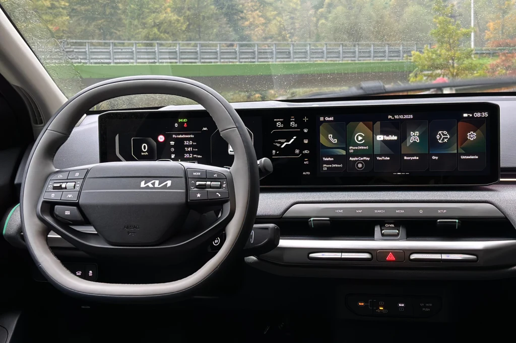 Koło dwuramiennej kierownicy skutecznie zasłania panel klimatyzacji kierowcy. Nowoczesna deska rozdzielcza samochodu marki Kia z cyfrowym wyświetlaczem, ekranem do obsługi multimediów, kierownicą z logo producenta oraz panelami sterowania klimatyzacją i przyciskami funkcyjnymi.