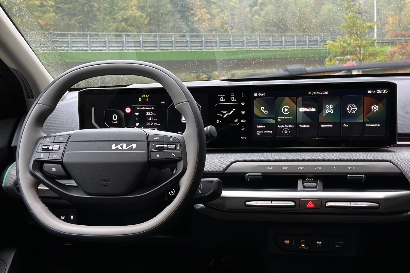 Koło dwuramiennej kierownicy skutecznie zasłania panel klimatyzacji kierowcy. Nowoczesna deska rozdzielcza samochodu marki Kia z cyfrowym wyświetlaczem, ekranem do obsługi multimediów, kierownicą z logo producenta oraz panelami sterowania klimatyzacją i przyciskami funkcyjnymi.