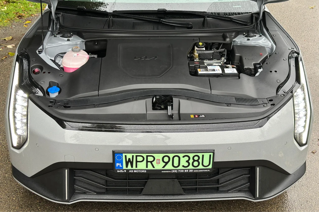 Pod maską nie ma "frunku". Jednostka elektryczna generuje moc 204 KM. Widok na komorę silnika nowoczesnego samochodu elektrycznego, gdzie w miejscu tradycyjnego silnika znajduje się schowek oraz elementy serwisowe takie jak akumulator i pojemnik na płyn chłodniczy, z przodu widoczna tablica rejestracyjna.