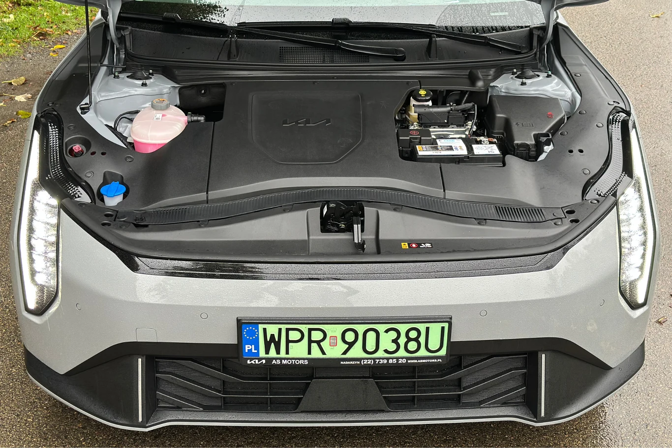 Pod maską nie ma "frunku". Jednostka elektryczna generuje moc 204 KM. Widok na komorę silnika nowoczesnego samochodu elektrycznego, gdzie w miejscu tradycyjnego silnika znajduje się schowek oraz elementy serwisowe takie jak akumulator i pojemnik na płyn chłodniczy, z przodu widoczna tablica rejestracyjna.