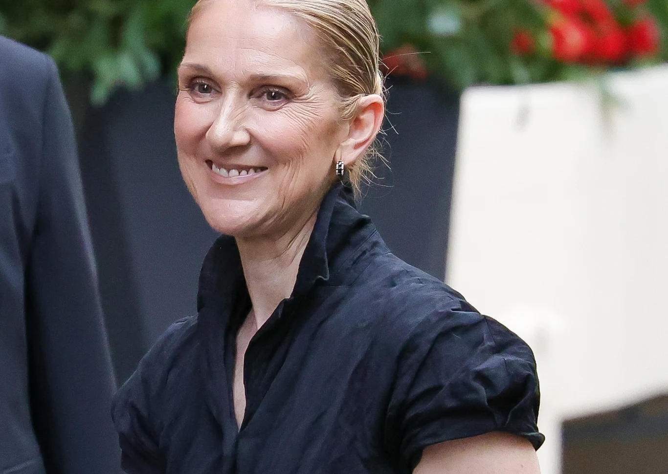 Celine Dion poruszyła fanów Uśmiechnięta kobieta z zaczesanymi do tyłu blond włosami, ubrana w elegancką czarną bluzkę, stoi przed rozmytym tłem z zielenią i czerwonymi kwiatami.