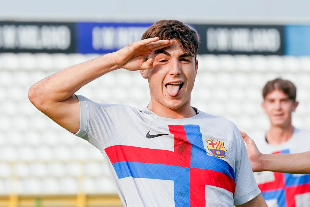 Victor Barbera w drużynie FC Barcelony U19 Młody piłkarz w białej koszulce FC Barcelony z krzyżem w barwach klubu wykonuje gest salutowania z wystawionym językiem, w tle rozmazany drugi zawodnik oraz trybuny stadionu.