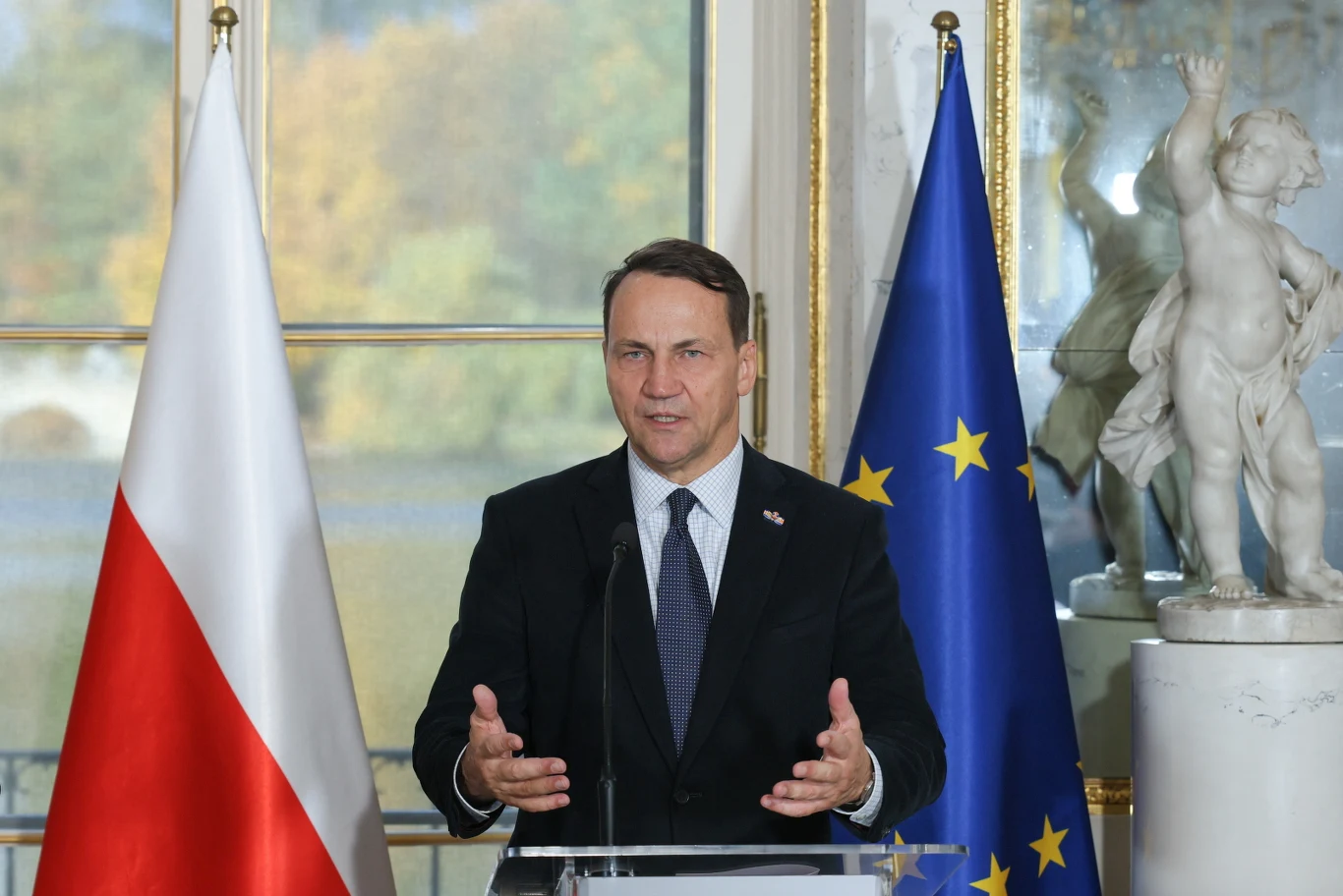 Szef MSZ Radosław Sikorski Mężczyzna w garniturze przemawia przy mównicy, za nim flaga Polski i flaga Unii Europejskiej, w tle biały posąg oraz widok na ogród przez duże okno.