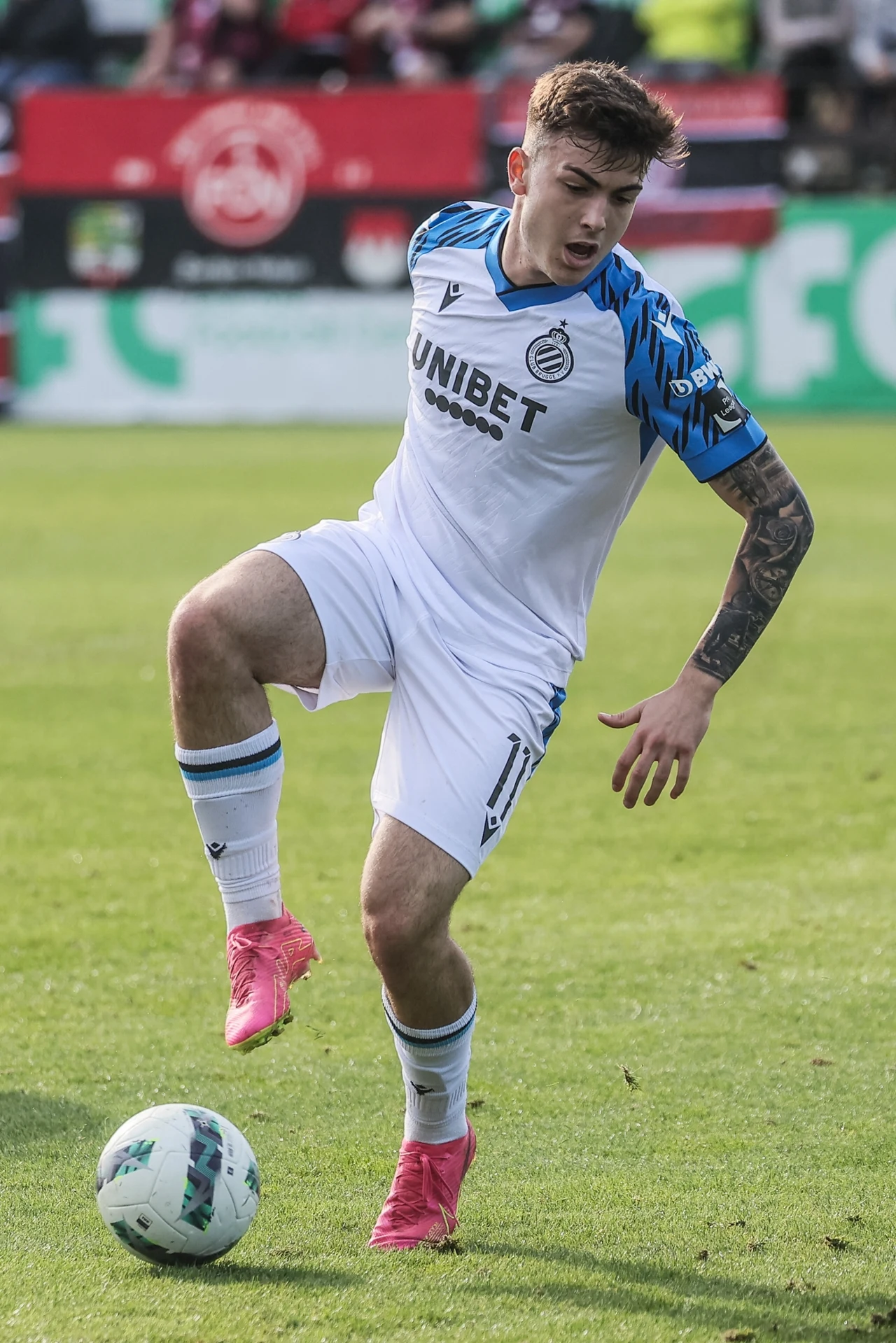 Victor Barbera w Club Brugge Piłkarz w białym stroju z niebieskimi detalami kontroluje piłkę na murawie podczas meczu, w tle widoczne trybuny i kibice.