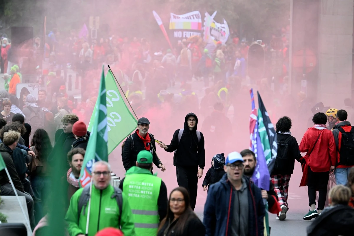 Wielotysięczne protesty w Belgii. Interweniowała policja Tłum ludzi maszerujących ulicą podczas demonstracji, z przodu widoczne flagi i transparenty, w tle unosi się różowy dym, część uczestników ubrana w jaskrawe kamizelki i kaptury.