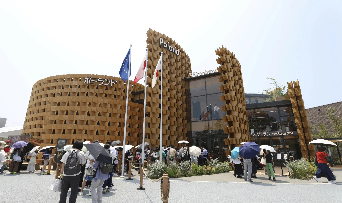 Wielki sukces Polski na Expo 2025 w Osace Nowoczesny budynek z charakterystyczną fasadą z drewnianych elementów, oznaczony napisem Poland, przed nim tłum ludzi z parasolami w słoneczny dzień, na pierwszym planie trzy flagi na masztach.