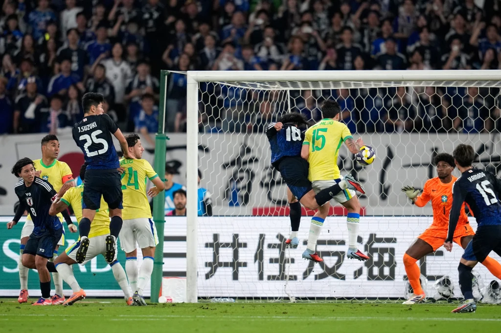 Kadr z meczy Brazylia - Japonia Piłkarze w ciemnoniebieskich i żółtych strojach walczą o piłkę blisko bramki, jeden z zawodników próbując zdobyć gola uderza głową, bramkarz i obrońcy starają się zablokować zagrożenie, tłum kibiców obserwuje sytuację z trybun