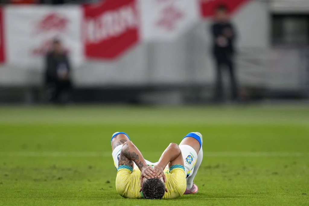 Kadr z meczu Brazylia - Japonia Piłkarz leżący na murawie boiska ze splecionymi dłońmi za głową, ubrany w żółtą koszulkę i niebieskie spodenki, w tle rozmazane postacie oraz biało-czerwone flagi na trybunach.