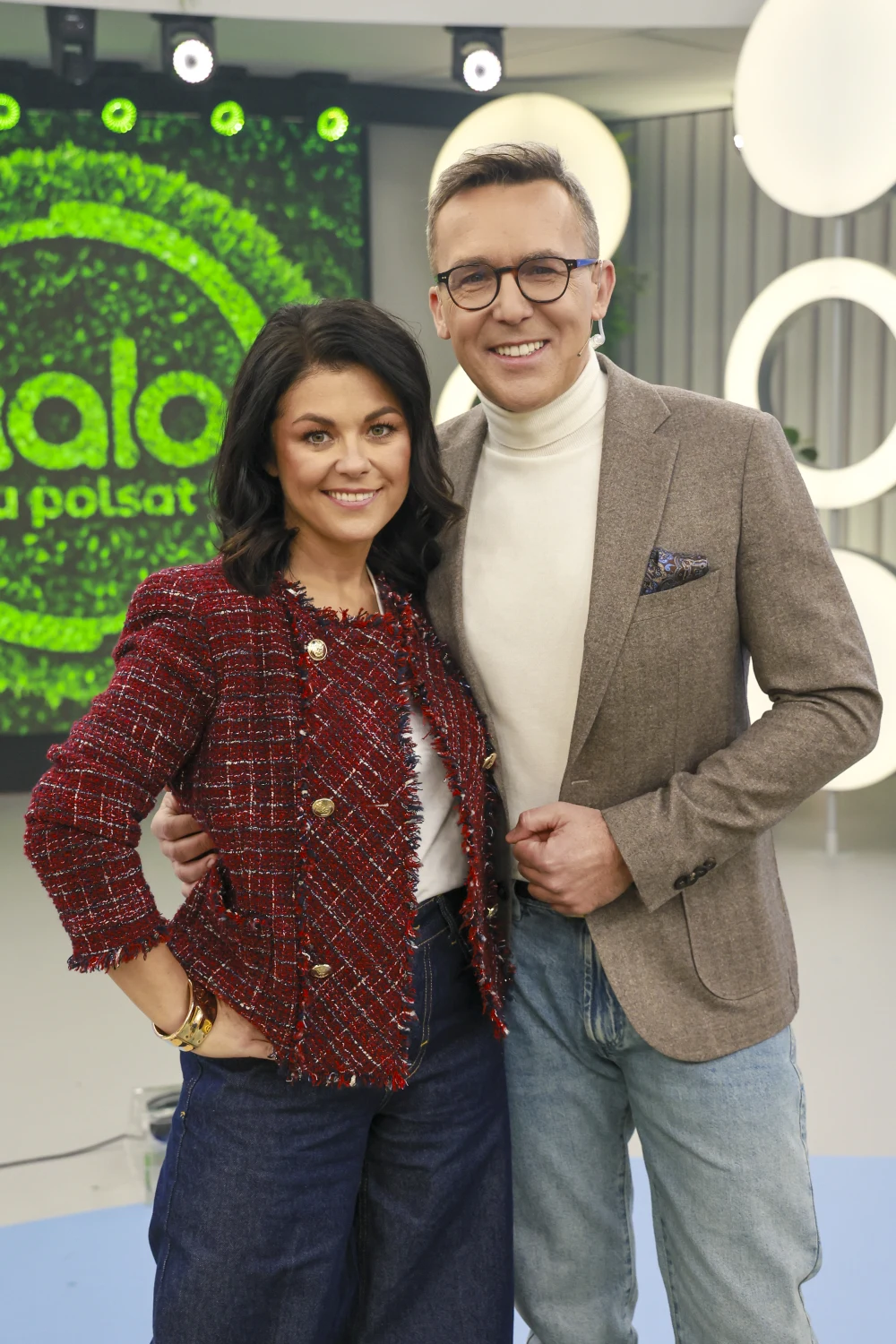 Katarzyna Cichopek w tweedowej marynarce Para dorosłych osób ubranych elegancko stoi razem w studiu telewizyjnym, w tle widoczne logo programu na zielonym ekranie.