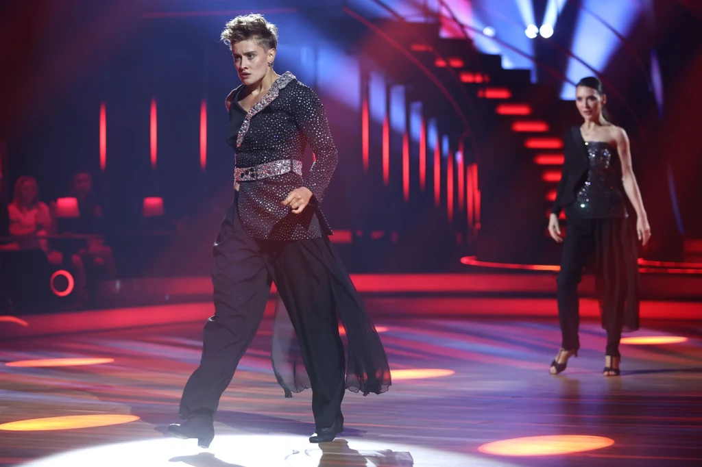 Katarzyna Zillmann Dynamiczna scena na parkiecie podczas występu tanecznego, na pierwszym planie mężczyzna w efektownym stroju, w tle kobieta, kolorowe światła i teatralna oprawa podkreślają atmosferę show