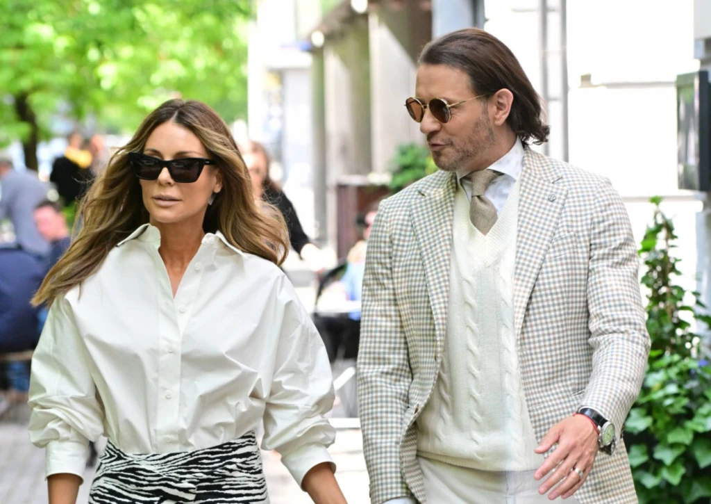 Małgorzata Rozenek, Radosław Majdan Elegancko ubrana kobieta i mężczyzna spacerują razem chodnikiem wśród zieleni, oboje mają okulary przeciwsłoneczne, wyglądają na zrelaksowanych i zamieniają ze sobą słowa.