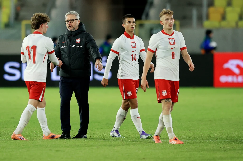 Jerzy Brzęczek i reprezentanci Polski do lat 21 Trener w czarnej kurtce rozmawiający z trzema młodymi piłkarzami ubranymi w białe koszulki i czerwone spodenki na boisku podczas meczu piłki nożnej.