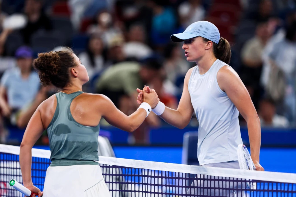 Jasmine Paolini i Iga Świątek Dwie tenisistki gratulujące sobie po meczu przy siatce na korcie tenisowym, obie ubrane w sportowe stroje, wokół nich widzowie na trybunach.