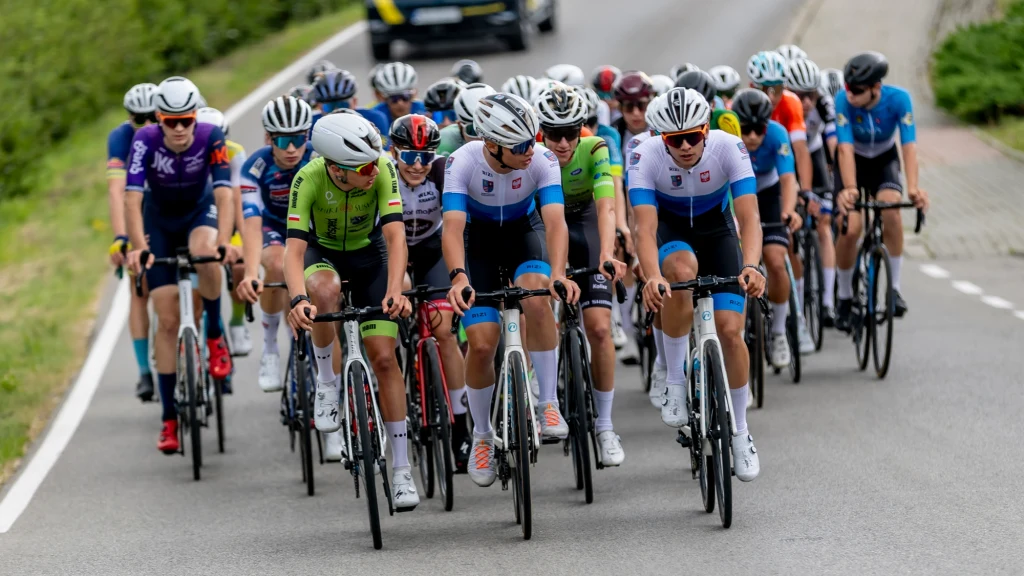 Grupa kolarzy w profesjonalnych strojach jedzie zwartą kolumną szosą podczas wyścigu, z tyłu widoczny samochód techniczny, zawodnicy skupieni i dynamiczni, otoczenie stanowi zielona roślinność po bokach drogi.