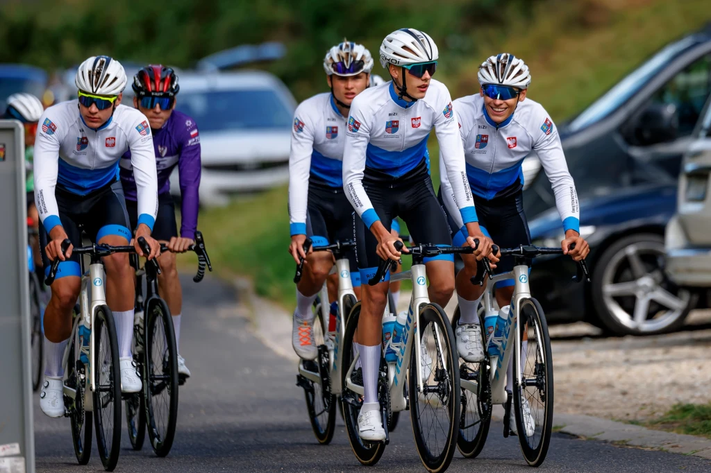 Grupa kolarzy w profesjonalnych strojach jedzie wspólnie szosą, wszyscy mają na sobie białe kaski i okulary sportowe, w tle widoczne są zaparkowane samochody oraz rozmyta przestrzeń zielona.