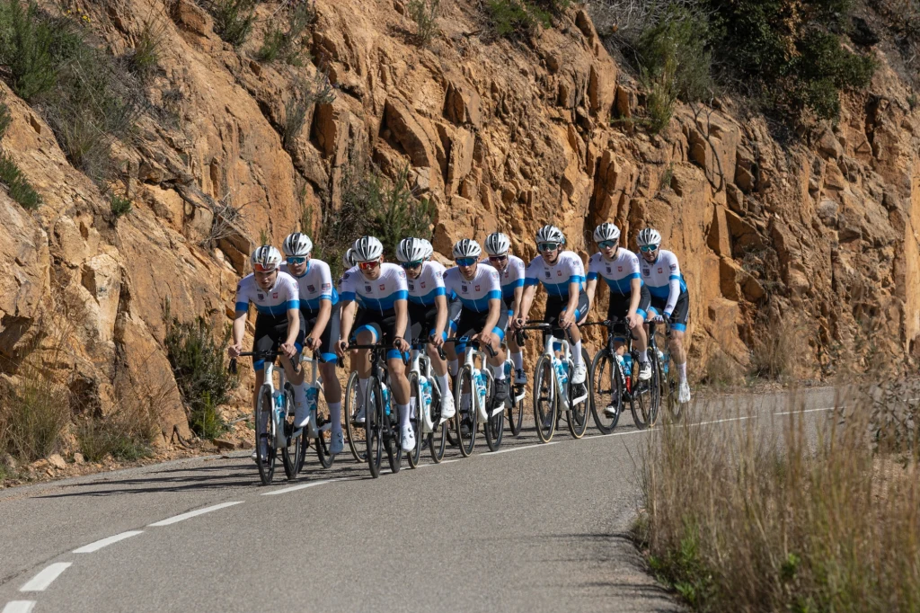 Grupa kolarzy w niebiesko-białych strojach jedzie w zwartej formacji po asfaltowej drodze wśród skalistych zboczy.