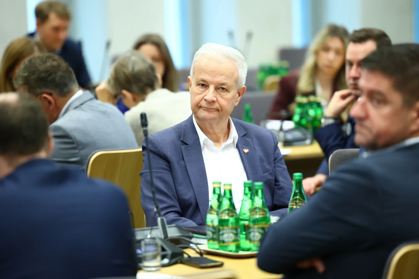 Starszy mężczyzna w garniturze siedzący przy stole konferencyjnym pośród innych osób, na stole stoją butelki zielonej wody mineralnej, w tle widać uczestników spotkania skupionych na rozmowie.