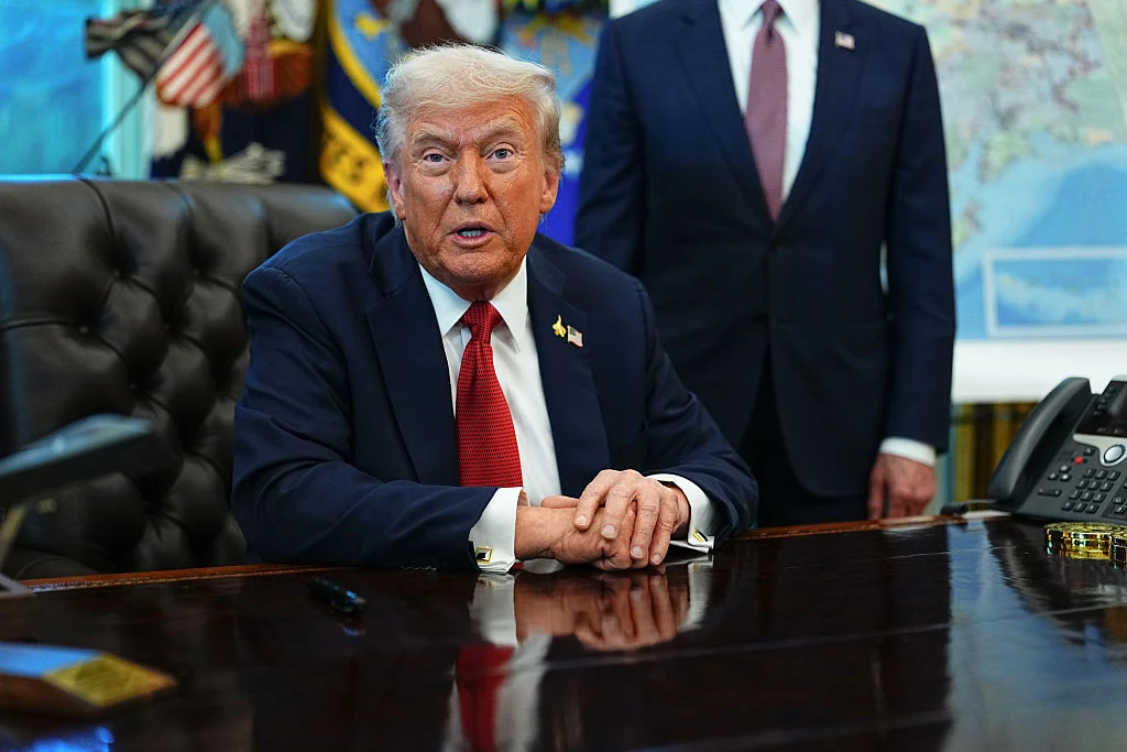 Donald Trump Mężczyzna w garniturze siedzi przy dużym biurku w luksusowym gabinecie, obok stoi inny mężczyzna w garniturze, w tle widoczne są flagi i fragment mapy.