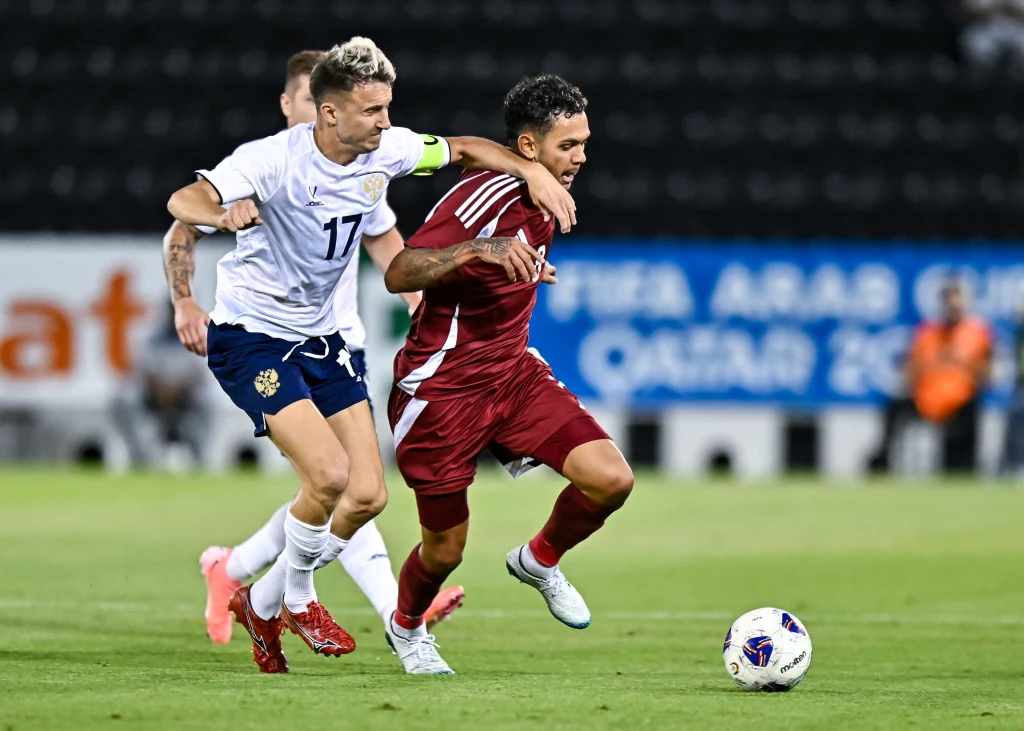 Dwóch piłkarzy ściga się o piłkę podczas meczu na stadionie, zawodnik w białej koszulce próbuje zatrzymać rywala w bordowym stroju, w tle widoczna murawa i rozmyte trybuny.