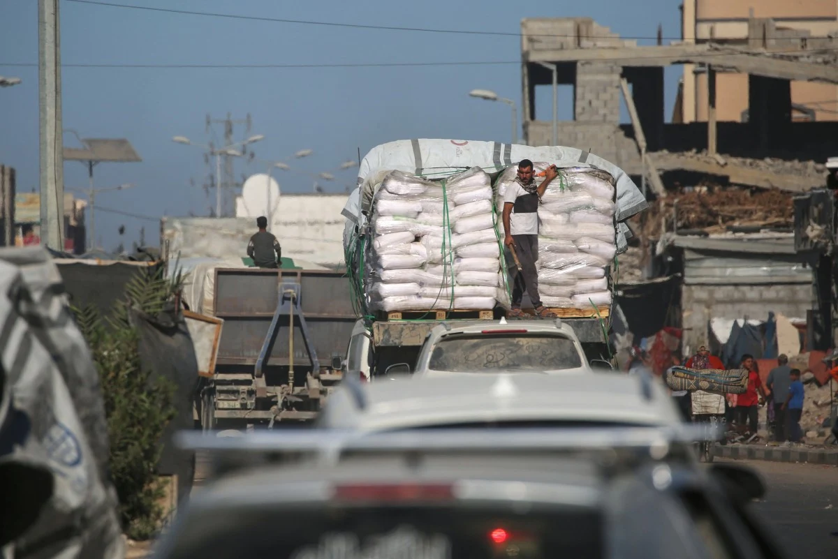 Pomoc humanitarna transportowana w Strefie Gazy Ciężarówka załadowana workami z pomocą humanitarną, na której stoi mężczyzna, porusza się ulicą wśród zniszczonych budynków i innych pojazdów, widoczne są ślady konfliktu oraz wyraźne zniszczenia infrastruktury.