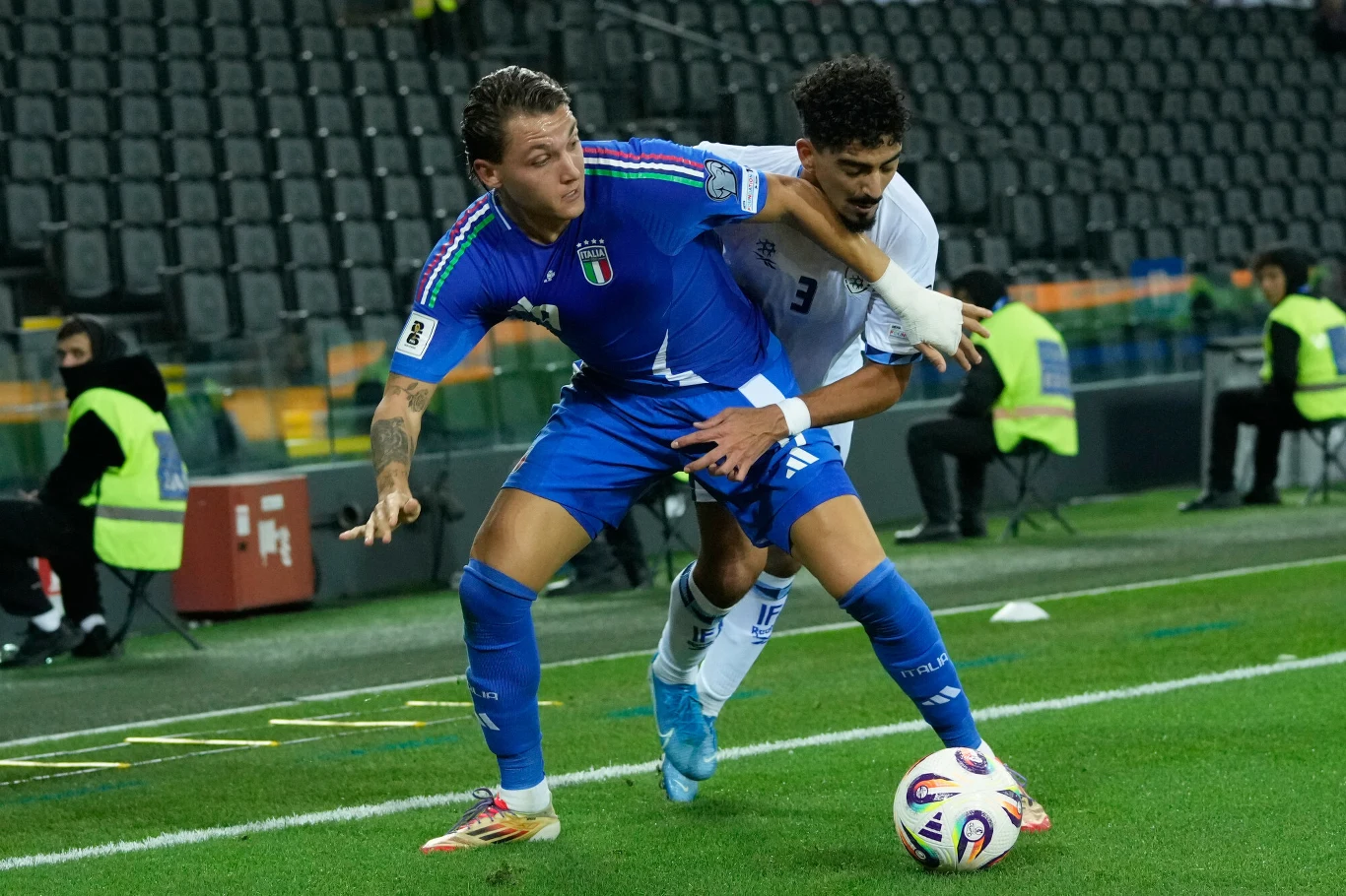 Dwóch piłkarzy rywalizuje o piłkę przy linii bocznej boiska, jeden ubrany w niebieski strój, drugi w biały, na trybunach widoczni są nieliczni obserwatorzy w kamizelkach odblaskowych.