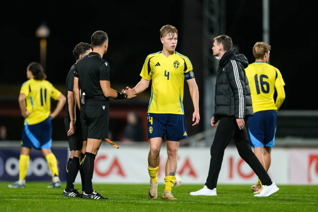 Zawodnik w żółtej koszulce z numerem 4 ściska dłoń sędziemu na boisku piłkarskim, wokół znajdują się inni piłkarze oraz trener w dresie, tło rozmyte, atmosfera po meczu.
