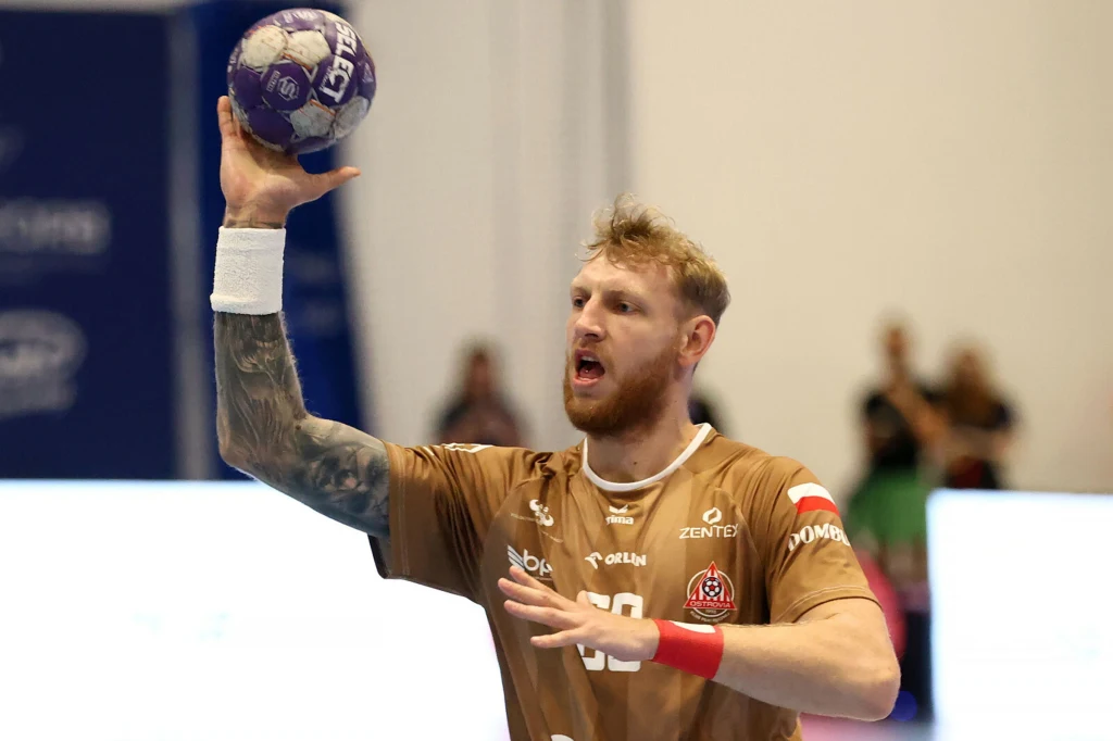 Ksawery Gajek, Re-Bud Ostrovia Ostrów Wlkp. Sportowiec w brązowej koszulce trzymający piłkę do piłki ręcznej przygotowuje się do rzutu na tle rozmazanej publiczności i neutralnego tła.