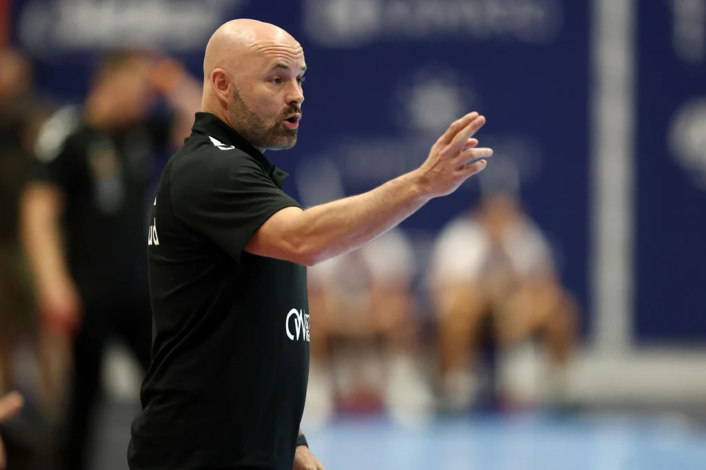 Kim Rasmussen, trener Ostrovii Mężczyzna w sportowej odzieży gestykuluje podczas wydarzenia sportowego, na tle rozmytych sylwetek osób widocznych w tle oraz niebieskiego tła z logotypami