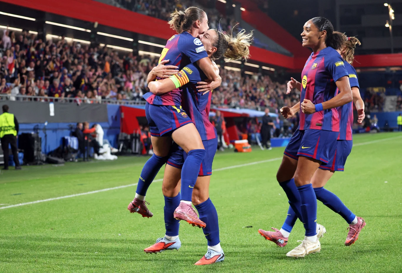 Piłkarki drużyny FC Barcelona świętują zdobycie bramki podczas meczu, jedna zawodniczka podnosi drugą w geście radości, reszta drużyny podbiega, aby dołączyć do świętowania, na trybunach tłum kibiców.