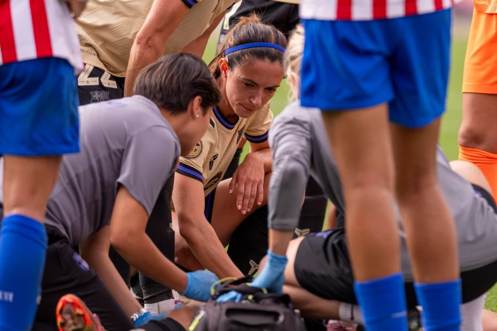 Kontuzja Ewy Pajor Kobieta w stroju sportowym kuca przy zawodniczce udzielającej pomocy medycznej przez członków sztabu podczas meczu piłkarskiego, wokół stoją inni sportowcy i ratownicy.