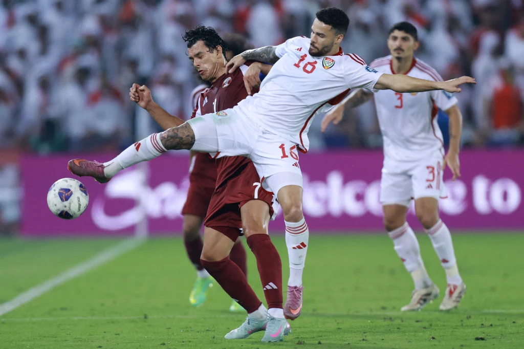 Dwóch piłkarzy w trakcie walki o piłkę na boisku, jeden z nich próbuje wykonać wślizg blokując przeciwnika, w tle rozmazana publiczność ubraną w białe stroje.