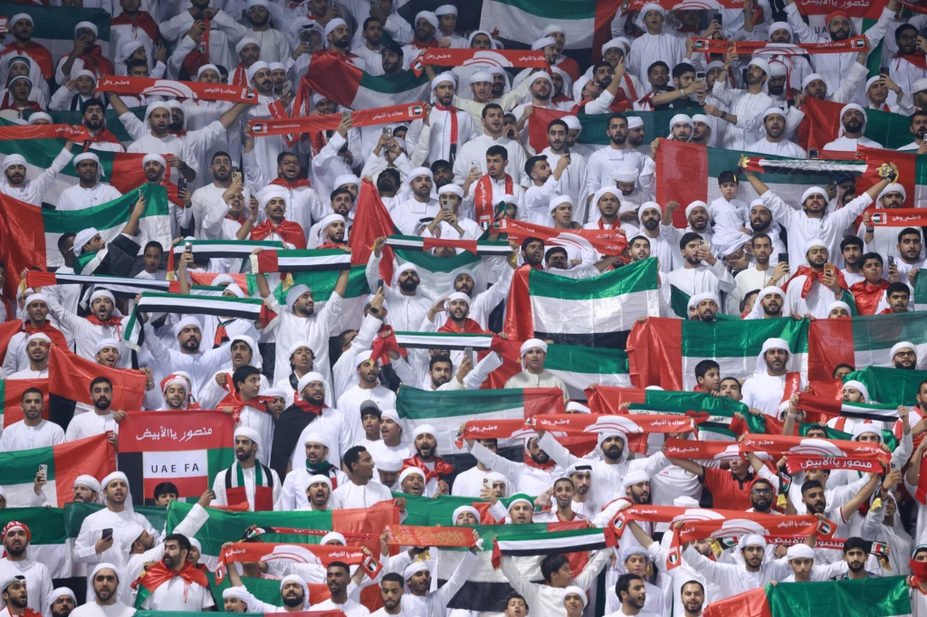 Tłum kibiców ubranych w tradycyjne białe stroje, trzymających flagi Zjednoczonych Emiratów Arabskich oraz czerwone i zielone szaliki, wyrażających wsparcie podczas wydarzenia sportowego.