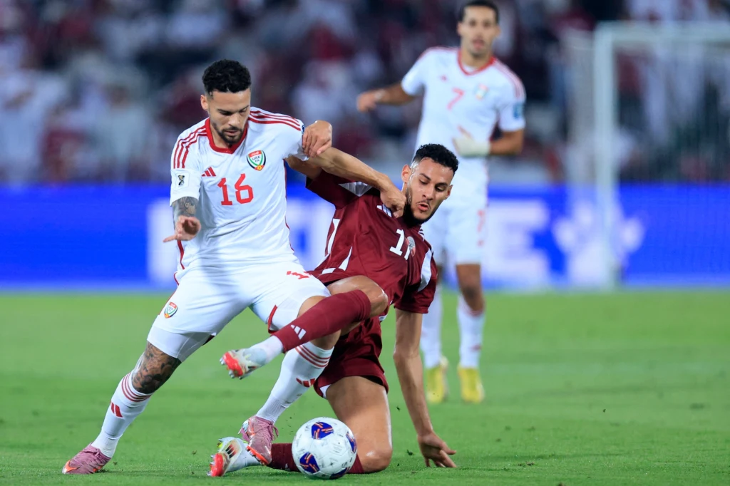 Dwóch piłkarzy walczy o piłkę na zielonym boisku, jeden w białym stroju z numerem 16, drugi w bordowym stroju z numerem 11, w tle rozmyci zawodnicy i widownia.