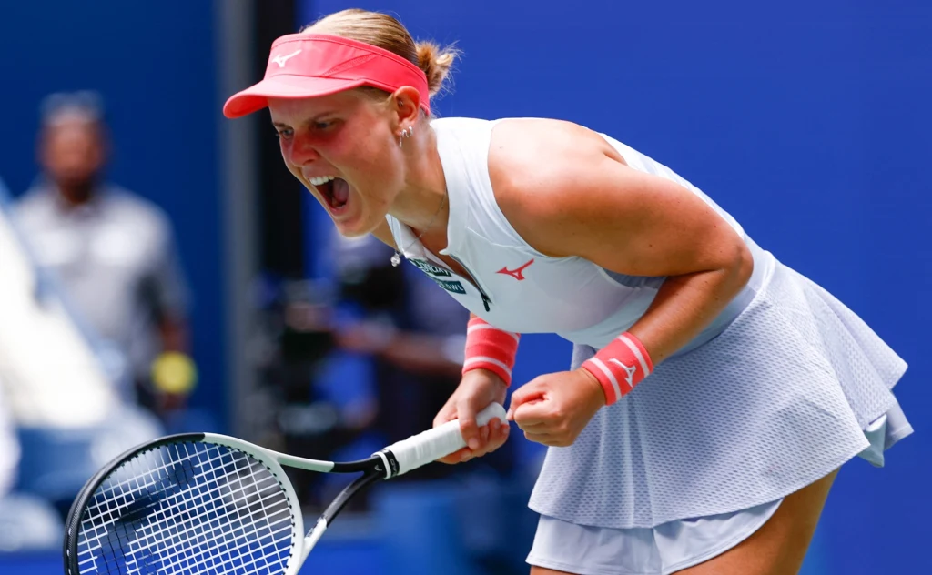 Suzan Lamens Tenisistka w białym stroju sportowym wykrzykuje z radości i zaciśniętymi pięściami trzyma rakietę tenisową, scena rozgrywa się na niebieskim korcie.