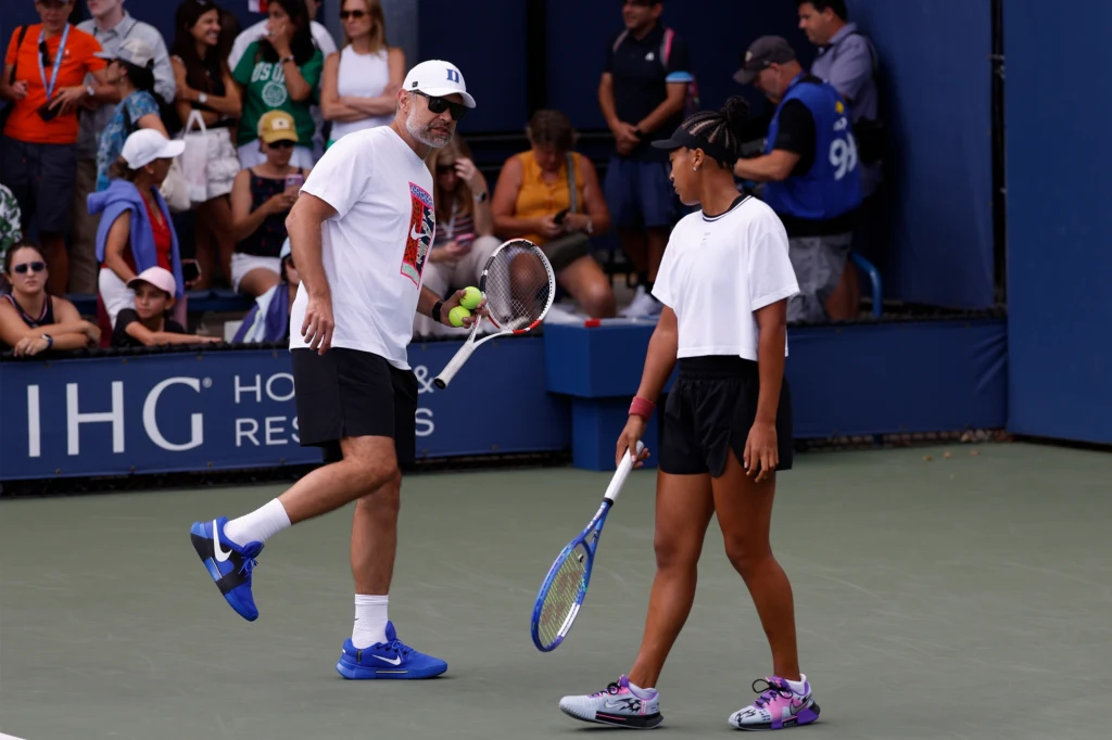 Tomasz Wiktorowski i Naomi Osaka Trener wymienia uwagi z tenisistką na korcie, wokół nich siedzą kibice obserwujący trening lub mecz, oboje trzymają rakiety tenisowe, a atmosfera skupienia i sportowej rywalizacji wyczuwalna jest wśród obecnych.