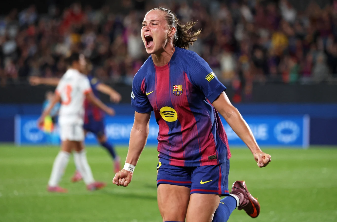 Ewa Pajor Piłkarka w stroju FC Barcelony celebruje zdobycie gola podczas meczu, z wyrazem wielkiej radości i uniesionymi pięściami, w tle znajduje się rozmazana sylwetka przeciwniczki oraz kibice na trybunach.