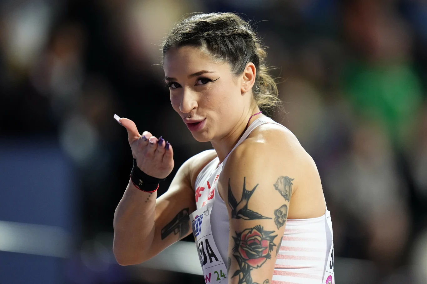 Uśmiechnięta kobieta sportowiec z tatuażami na ramionach, ubrana w sportowy strój, wykonuje gest pocałunku dłonią skierowany do widza, tło rozmyte z widownią.