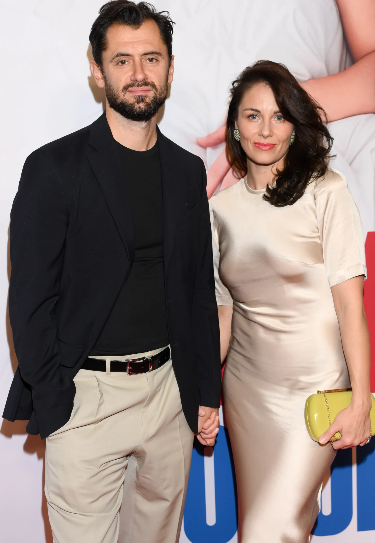 Bartlomiej Kotschedoff i Katarzyna Wajda na premierze filmowej Elegancko ubrany mężczyzna w czarnym t-shircie, kremowych spodniach i marynarce stoi obok kobiety w satynowej sukience w kolorze ecru, trzymającej żółtą kopertówkę. Trzymają się za ręce na tle jasnej ścianki z nadrukiem.