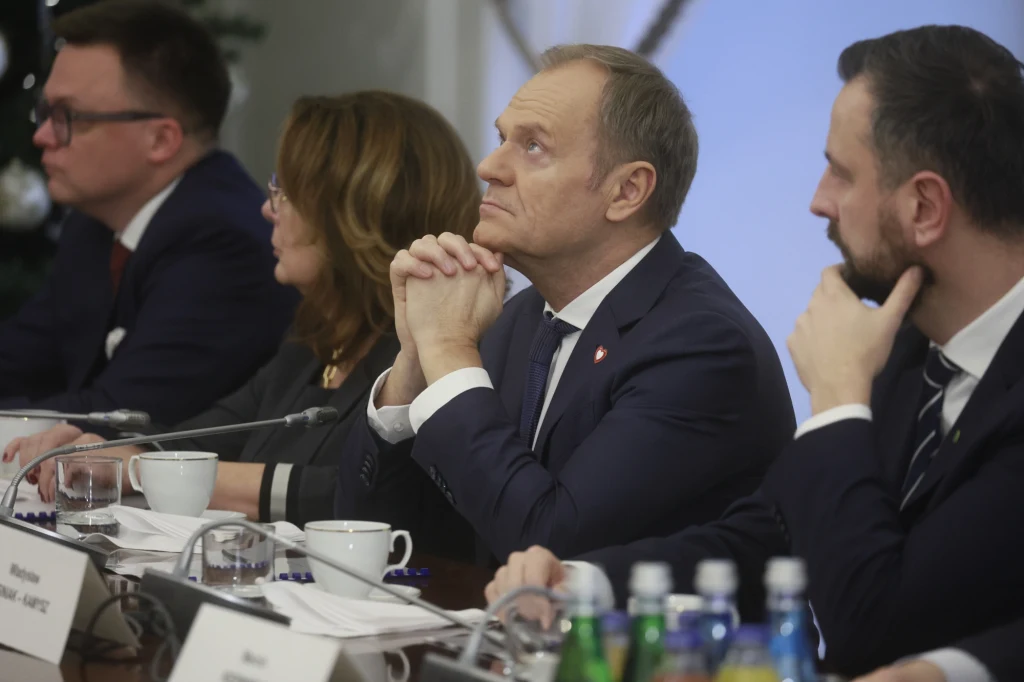 Grupa osób w garniturach i formalnych ubraniach siedzi przy stole konferencyjnym, skupiona na rozmowie lub prezentacji, przed nimi filiżanki oraz butelki z wodą mineralną.