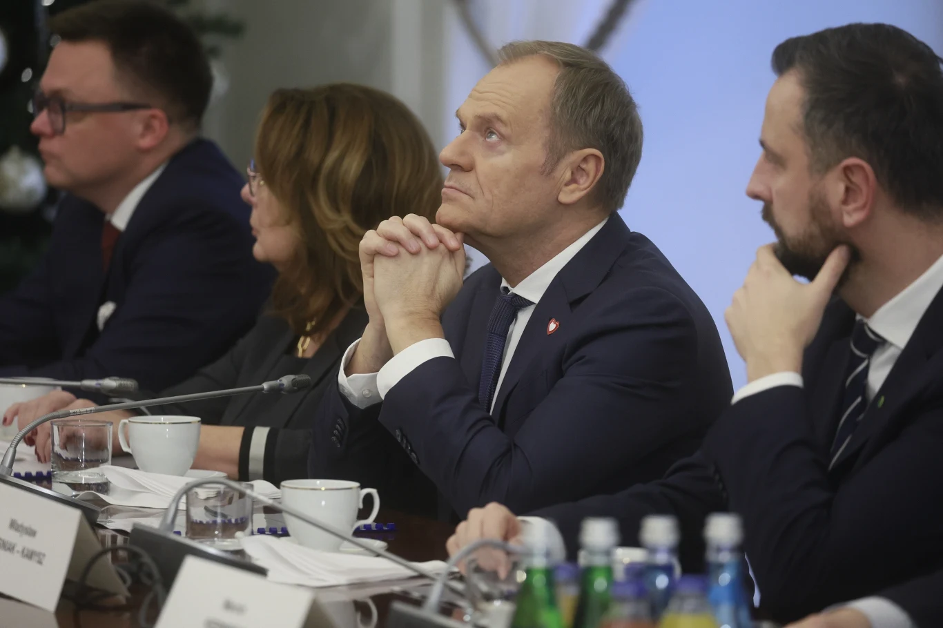 Grupa osób w garniturach i formalnych ubraniach siedzi przy stole konferencyjnym, skupiona na rozmowie lub prezentacji, przed nimi filiżanki oraz butelki z wodą mineralną.