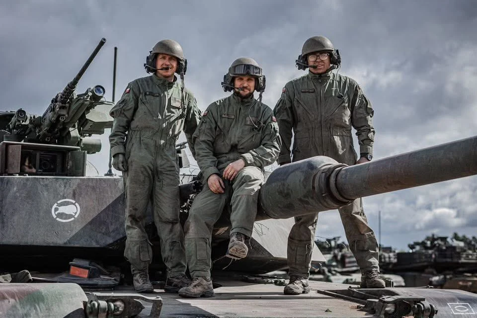 Załoganci czołgi M1A1 Abrams z 1. Warszawskiej Brygady Pancernej z widocznymi hełmofonami z czepcami balistycznymi Trzech żołnierzy w mundurach i kaskach stoi oraz siedzi na czołgu, wokół widoczne są inne pojazdy wojskowe na tle pochmurnego nieba.