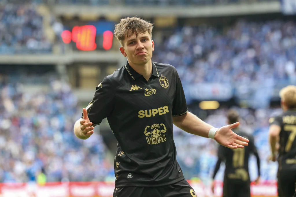 Młody piłkarz w czarnej koszulce z napisem SUPER oraz oznaczeniem klubu wykonuje charakterystyczny gest z rękami uniesionymi na boki, w tle rozmyte trybuny pełne kibiców.