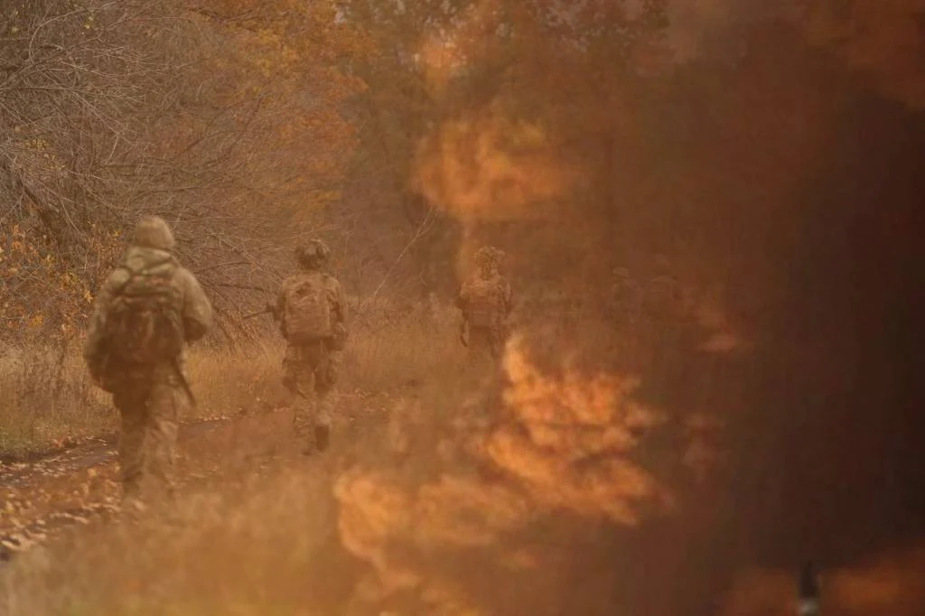 16 zbiorników paliwa zostało uszkodzonych w wyniku ukraińskiego ataku na Morski Terminal Naftowy Żołnierze w pełnym umundurowaniu przemierzają leśną ścieżkę, jakby podczas patrolu, z jesiennymi liśćmi w tle. Po prawej stronie widoczny rozmyty fragment ognia lub dymu, co sugeruje bliskość zagrożenia lub intensywne działania.