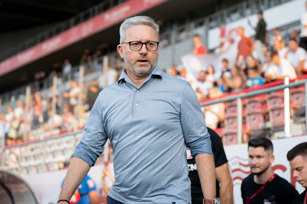 Mężczyzna w okularach i koszuli z krótkim rękawem na trybunach stadionu, w tle rozmyci kibice i elementy stadionu sportowego.