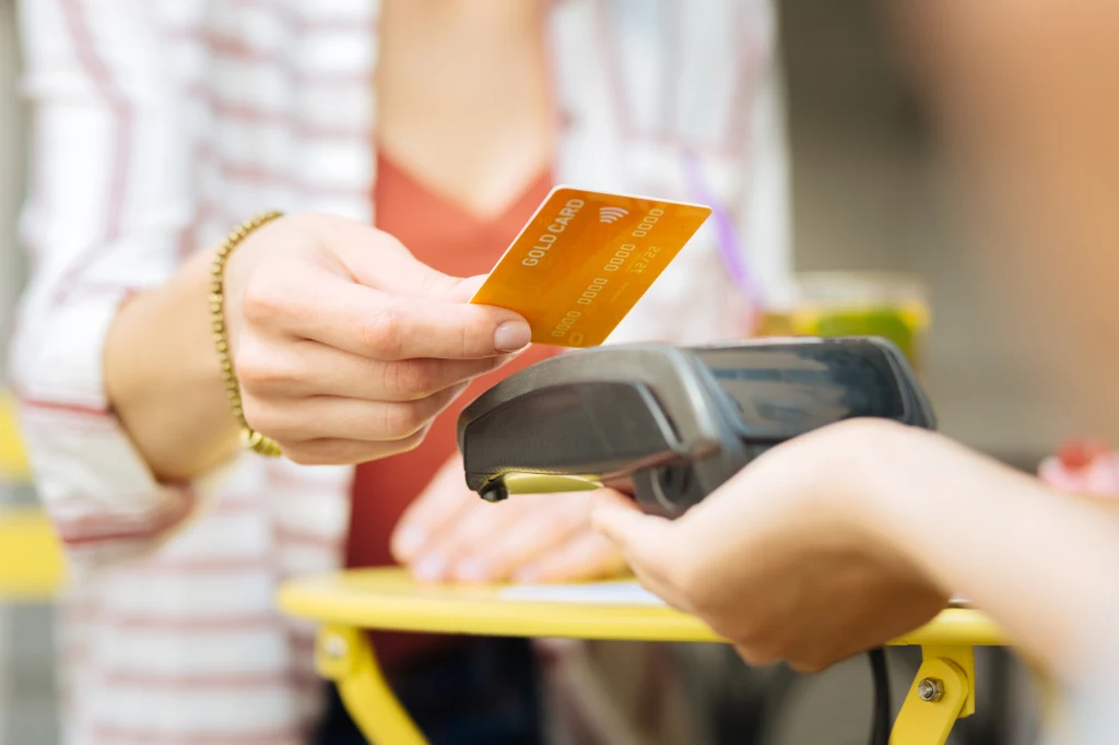 Niektórych wydatków nawet nie zauważymy - dopóki nie sprawdzimy dokładnego stanu konta Ręka trzymająca złotą kartę płatniczą zbliża kartę do terminala płatniczego w celu dokonania płatności bezdotykowej, w tle widoczne rozmyte elementy ubioru oraz blat stolika.