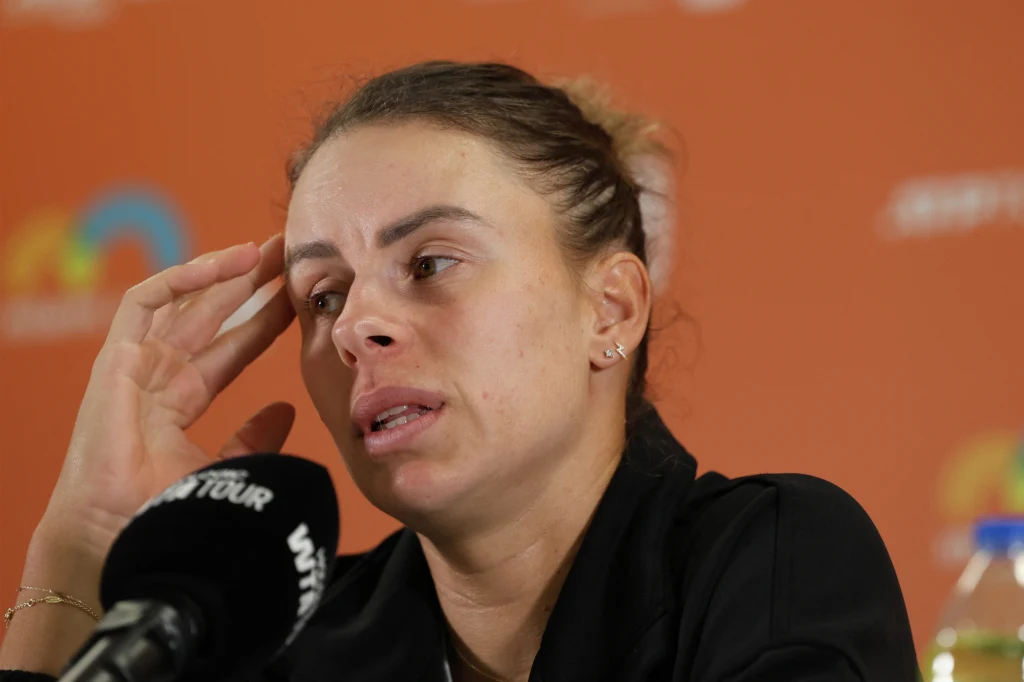 Magda Linette Kobieta z wyraźnym wyrazem zmęczenia lub zatroskania na twarzy, trzymająca dłoń przy skroni, siedzi przed mikrofonem na tle pomarańczowej ścianki z logotypami, sugerującej wydarzenie sportowe lub konferencję prasową.