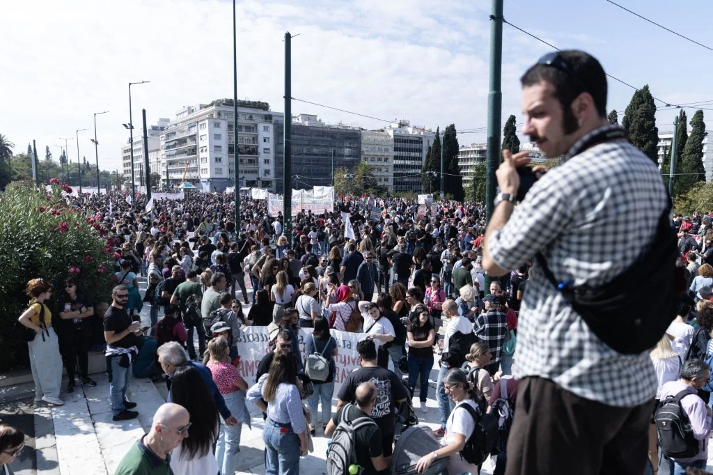 Tłum ludzi zgromadzonych podczas protestu na ulicy otoczonej wysokimi budynkami, część uczestników trzyma transparenty, w prawym przednim planie stoi mężczyzna z kamerą rejestrujący wydarzenia.